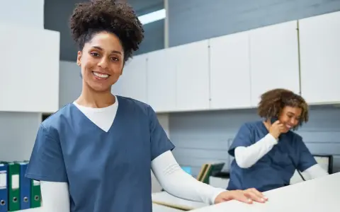 Une femme souriante en uniforme médical, prête pour sa formation secrétaire médicale en ligne. Une collègue au téléphone en arrière-plan.