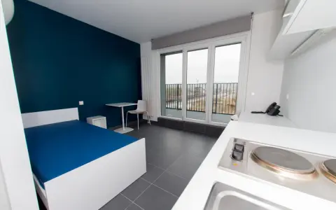 Chambre moderne pour étudiant à Luxembourg : lit bleu, bureau, kitchenette et balcon avec vue.