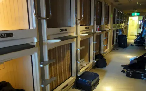 Couloir d'un hôtel capsule au Japon, avec des rangées de compartiments pour le **loyer au Japon**. Des valises sont posées au sol.