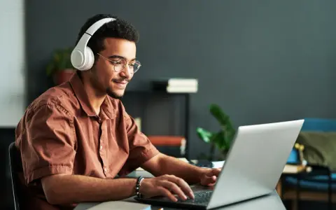 Jeune homme souriant avec des écouteurs, concentré sur son ordinateur portable. Il suit un master informatique à distance, profitant de la flexibilité de l'apprentissage en ligne.