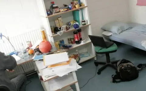 Bureau désordonné, lit simple, étagère remplie d'articles divers. Un ordinateur portable sur une chaise. Une scène typique pour trouver un logement en Belgique.