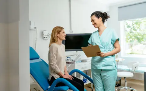 Une femme en blouse médicale parle à une patiente assise sur un fauteuil d'examen bleu.