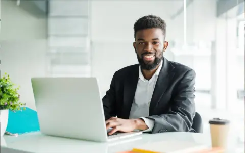 Un homme souriant travaille sur son ordinateur portable, peut-être pour son msc finance online. Il est dans un bureau moderne.