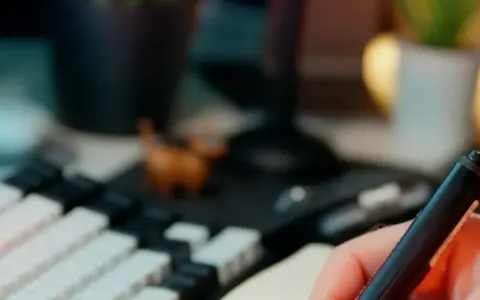 Main tenant une plume, prêt à étudier pour sa licence sociologie à distance. Un clavier mécanique et une petite figurine complètent le décor.