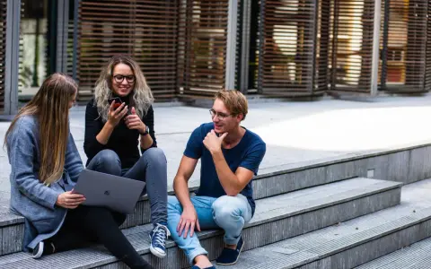 Trois jeunes discutent sur des marches, l'un regarde son téléphone, l'autre un ordinateur portable. Ils semblent parler du cout de la vie aux Pays-Bas.