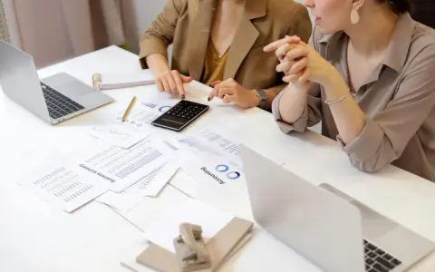 Deux femmes analysent des documents financiers et utilisent une calculatrice. Leurs mains pointent vers des graphiques, suggérant une discussion sur le pilotage de la performance, peut-être dans le cadre d'un msc audit à KEDGE.