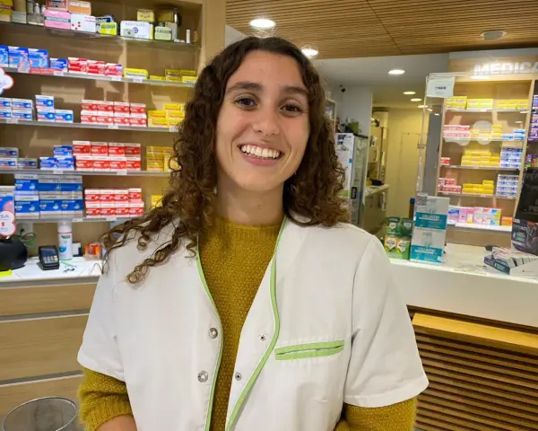 Une souriante préparatrice en pharmacie en Espagne, entourée de boîtes de médicaments sur les étagères.