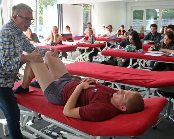 Un professeur pratique un examen de genou sur un étudiant allongé sur une table de massage rouge, dans une salle de classe d'une école de kinésithérapie suisse.