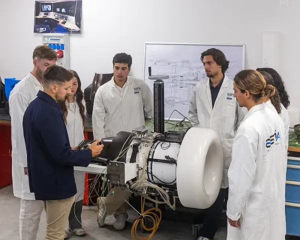 Des étudiants en blouse blanche observent un moteur complexe dans un laboratoire d'école d'ingénieur en Belgique.