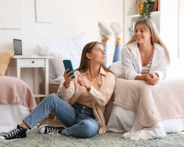 Deux étudiantes françaises discutent de leur aide au logement en Espagne, l'une sur le lit, l'autre assise au sol avec un téléphone.