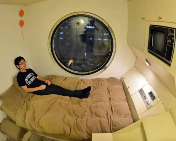 Un jeune homme se détend dans un lit, dans un petit logement étudiant à Tokyo. Une fenêtre ronde offre une vue nocturne sur la ville.
