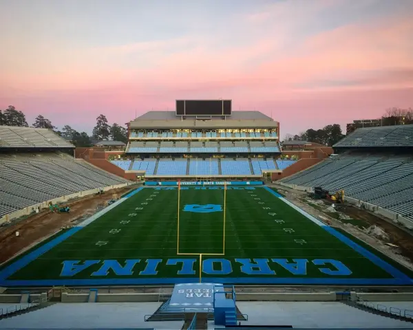 Stade de football vide de l'université de Caroline du Nord à Chapel Hill, avec des sièges bleus et le logo des Tar Heels. Le ciel rose du crépuscule surplombe le terrain.