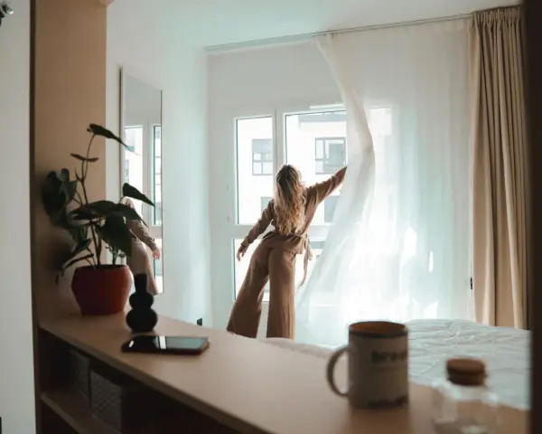 Une jeune femme aux cheveux blonds dénoue les rideaux d'une chambre lumineuse. Un aperçu de la vie étudiante à Genève, où le logement étudiant et son prix sont une préoccupation majeure.