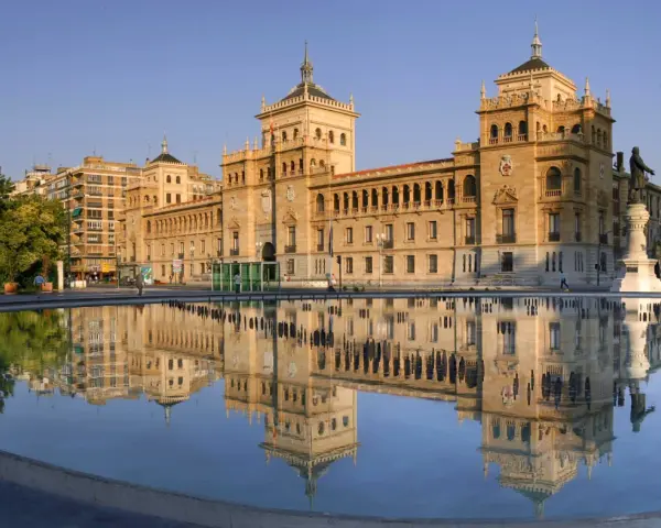 Le majestueux bâtiment du **universidad de valladolid** se reflète dans l'eau calme d'une fontaine, sous un ciel bleu clair.