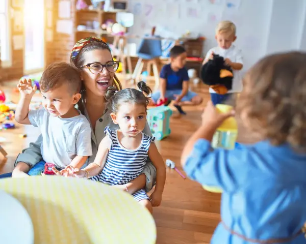 Une enseignante souriante, en formation **master petite enfance à distance**, interagit avec des enfants dans une salle de jeux colorée.