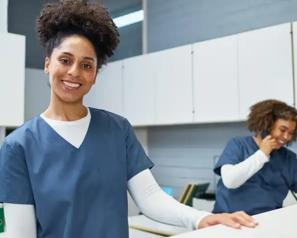 Une femme souriante en uniforme médical, prête pour sa formation secrétaire médicale en ligne. Une collègue au téléphone en arrière-plan.