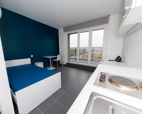 Chambre moderne pour étudiant à Luxembourg : lit bleu, bureau, kitchenette et balcon avec vue.