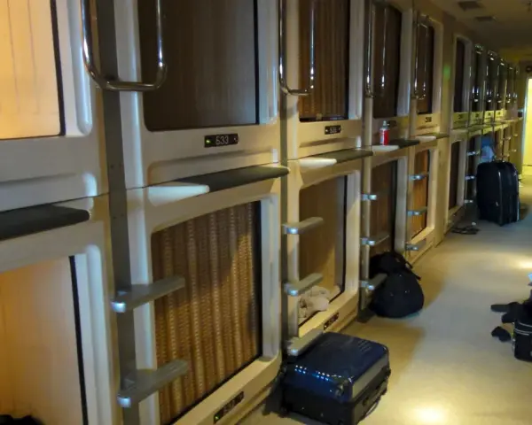Couloir d'un hôtel capsule au Japon, avec des rangées de compartiments pour le **loyer au Japon**. Des valises sont posées au sol.