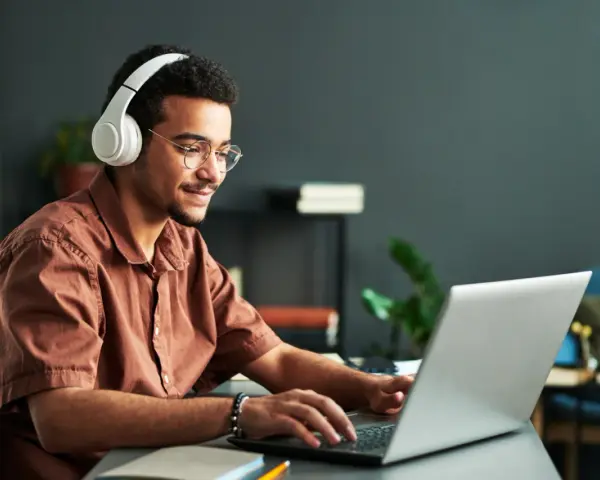 Jeune homme souriant avec des écouteurs, concentré sur son ordinateur portable. Il suit un master informatique à distance, profitant de la flexibilité de l'apprentissage en ligne.