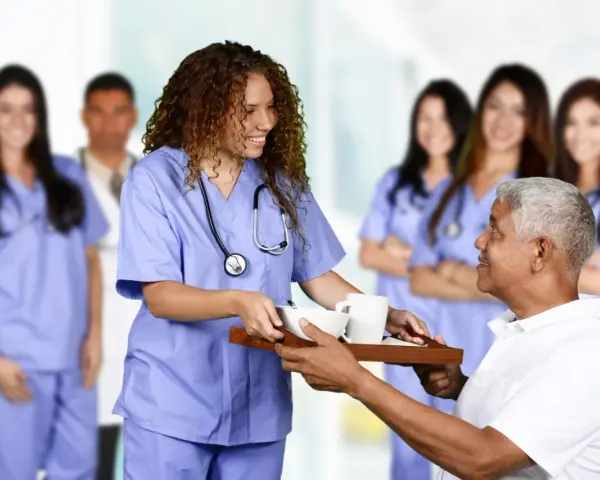 Une jeune femme en formation paramédicale sans bac, souriante, sert un plateau repas à un patient.