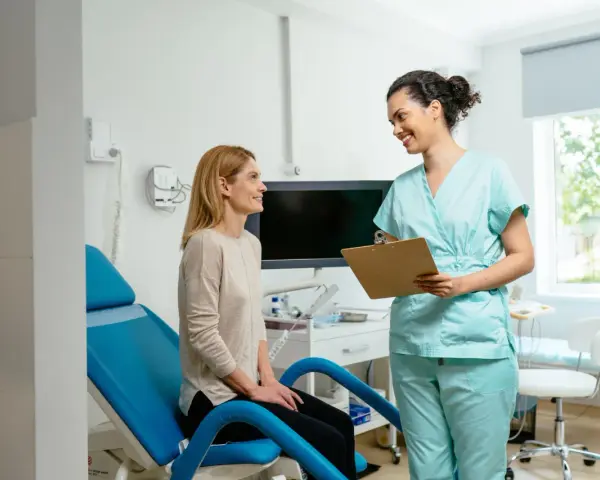 Une femme en blouse médicale parle à une patiente assise sur un fauteuil d'examen bleu.