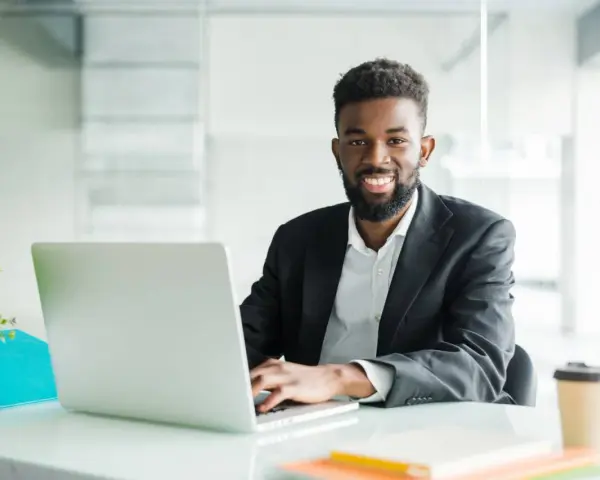 Un homme souriant travaille sur son ordinateur portable, peut-être pour son msc finance online. Il est dans un bureau moderne.