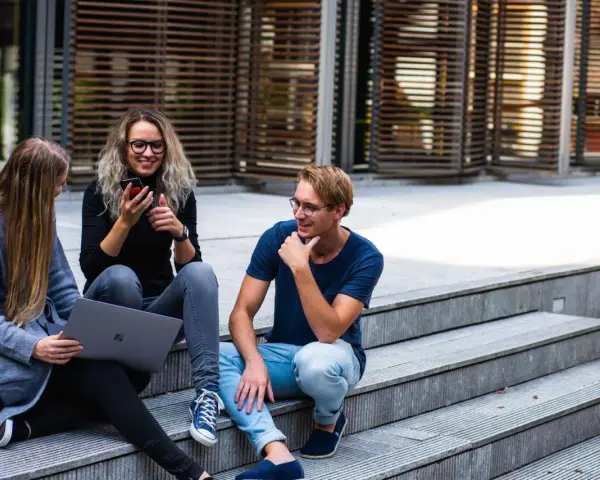 Trois jeunes discutent sur des marches, l'un regarde son téléphone, l'autre un ordinateur portable. Ils semblent parler du cout de la vie aux Pays-Bas.