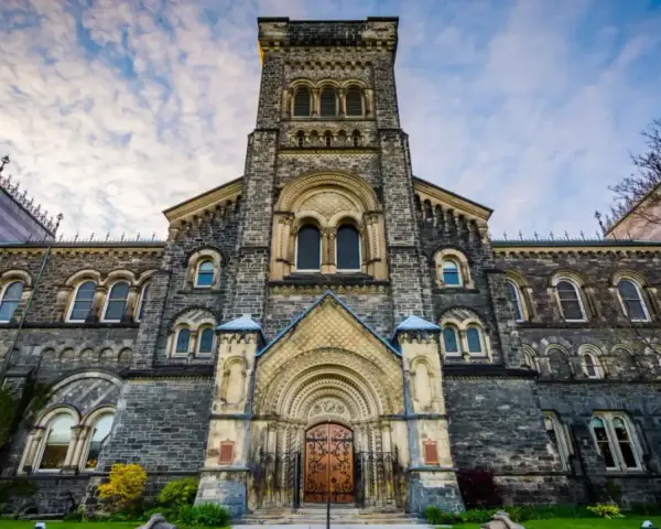 La façade imposante de l'université de Toronto, avec sa tour centrale et ses fenêtres cintrées, se dresse sous un ciel nuageux.