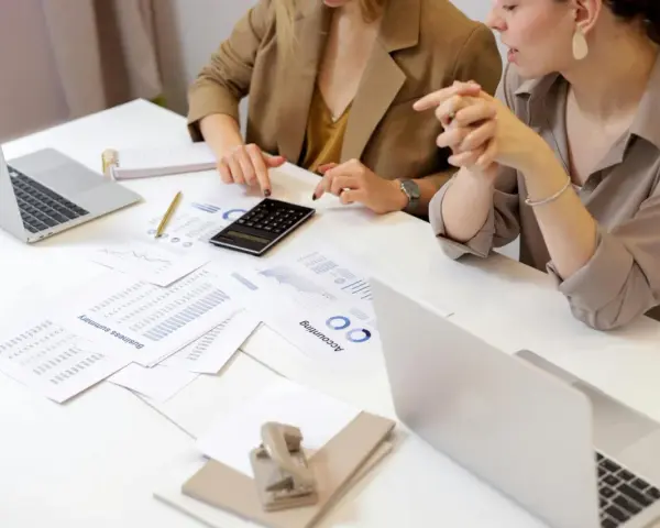 Deux femmes analysent des documents financiers et utilisent une calculatrice. Leurs mains pointent vers des graphiques, suggérant une discussion sur le pilotage de la performance, peut-être dans le cadre d'un msc audit à KEDGE.