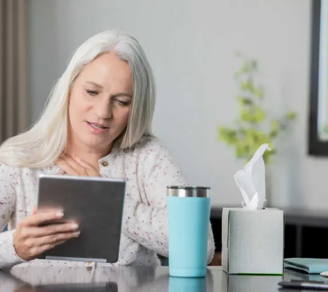 Femme aux cheveux gris suivant une formation orthophoniste à distance sur sa tablette, une tasse et des mouchoirs à proximité.