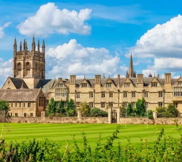 Magnifique vue sur l'université d'Oxford, avec ses bâtiments historiques en pierre, ses tours imposantes et son vaste terrain de jeu verdoyant sous un ciel bleu parsemé de nuages.