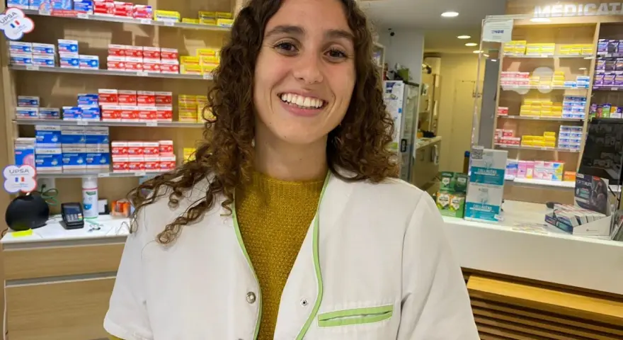 Une souriante préparatrice en pharmacie en Espagne, entourée de boîtes de médicaments sur les étagères.