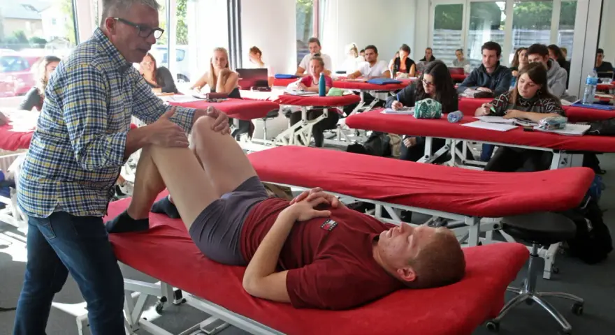 Un professeur pratique un examen de genou sur un étudiant allongé sur une table de massage rouge, dans une salle de classe d'une école de kinésithérapie suisse.