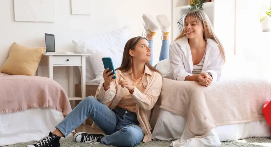 Deux étudiantes françaises discutent de leur aide au logement en Espagne, l'une sur le lit, l'autre assise au sol avec un téléphone.