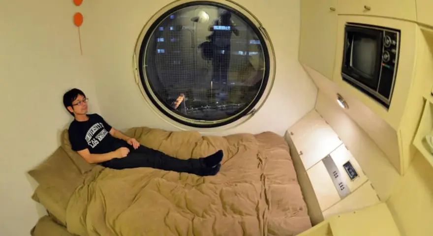 Un jeune homme se détend dans un lit, dans un petit logement étudiant à Tokyo. Une fenêtre ronde offre une vue nocturne sur la ville.