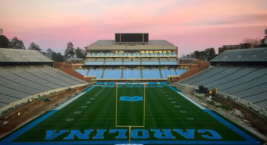 Stade de football vide de l'université de Caroline du Nord à Chapel Hill, avec des sièges bleus et le logo des Tar Heels. Le ciel rose du crépuscule surplombe le terrain.