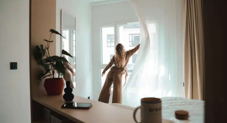 Une jeune femme aux cheveux blonds dénoue les rideaux d'une chambre lumineuse. Un aperçu de la vie étudiante à Genève, où le logement étudiant et son prix sont une préoccupation majeure.