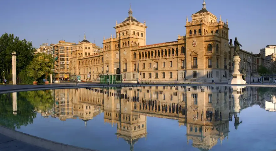 Le majestueux bâtiment du **universidad de valladolid** se reflète dans l'eau calme d'une fontaine, sous un ciel bleu clair.