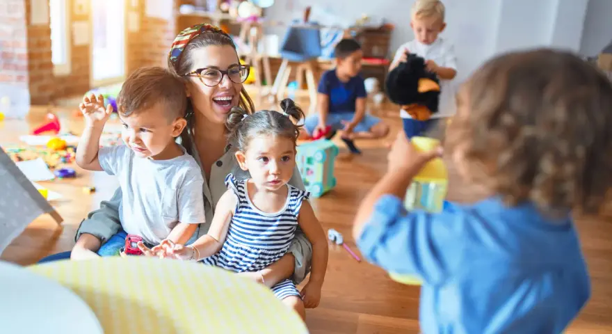Une enseignante souriante, en formation **master petite enfance à distance**, interagit avec des enfants dans une salle de jeux colorée.