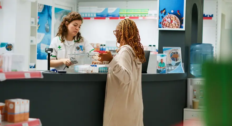Une pharmacienne conseille une cliente. Une affiche sur la formation préparateur en pharmacie à distance est visible.