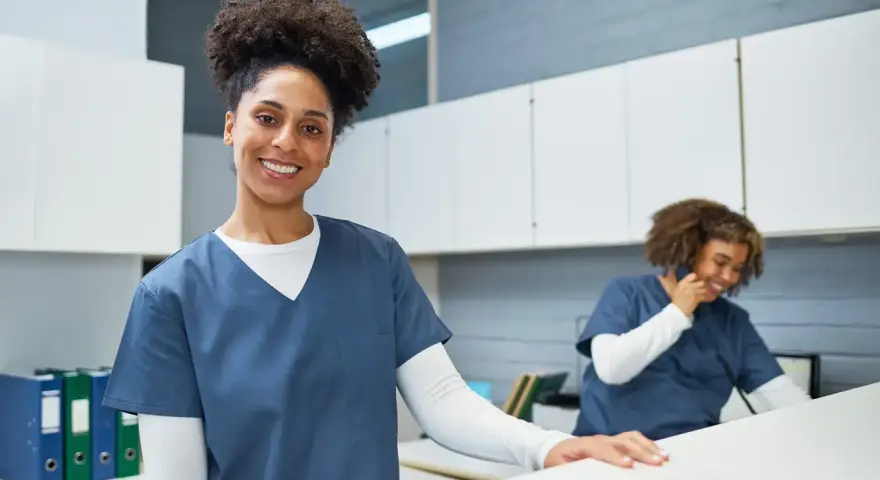 Une femme souriante en uniforme médical, prête pour sa formation secrétaire médicale en ligne. Une collègue au téléphone en arrière-plan.