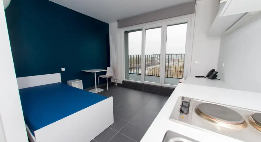 Chambre moderne pour étudiant à Luxembourg : lit bleu, bureau, kitchenette et balcon avec vue.