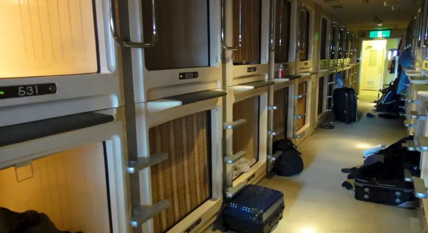 Couloir d'un hôtel capsule au Japon, avec des rangées de compartiments pour le **loyer au Japon**. Des valises sont posées au sol.