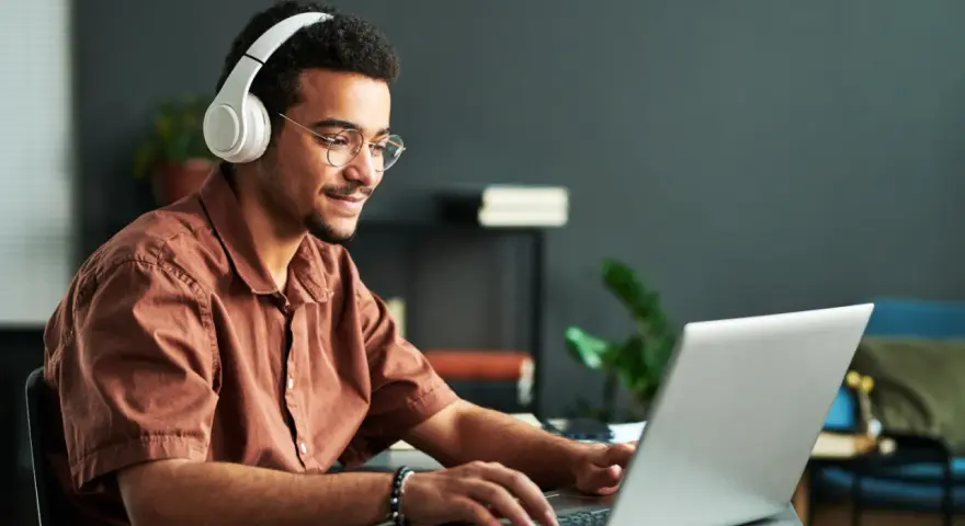Jeune homme souriant avec des écouteurs, concentré sur son ordinateur portable. Il suit un master informatique à distance, profitant de la flexibilité de l'apprentissage en ligne.