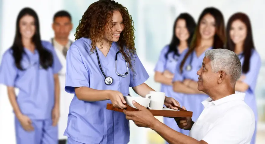 Une jeune femme en formation paramédicale sans bac, souriante, sert un plateau repas à un patient.
