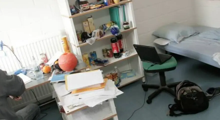 Bureau désordonné, lit simple, étagère remplie d'articles divers. Un ordinateur portable sur une chaise. Une scène typique pour trouver un logement en Belgique.