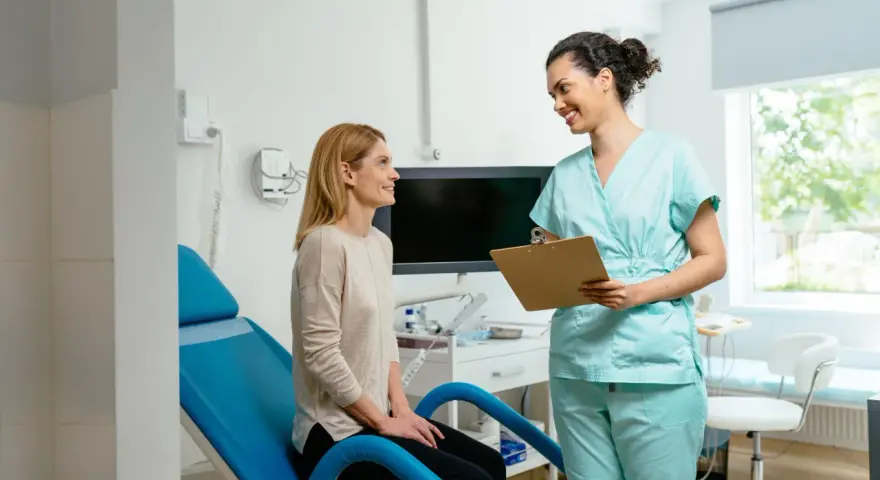 Une femme en blouse médicale parle à une patiente assise sur un fauteuil d'examen bleu.