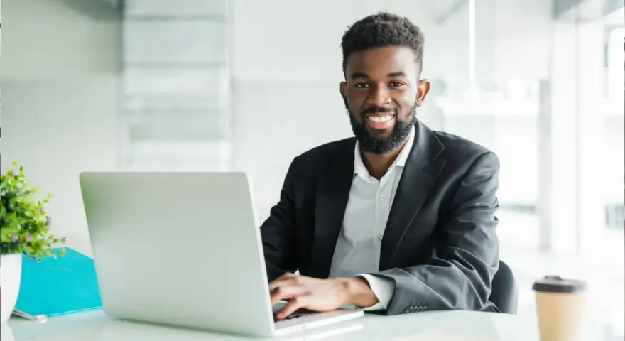 Un homme souriant travaille sur son ordinateur portable, peut-être pour son msc finance online. Il est dans un bureau moderne.