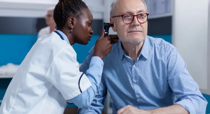 Une jeune femme en formation BTS Audioprothésiste examine l'oreille d'un patient âgé avec un otoscope.