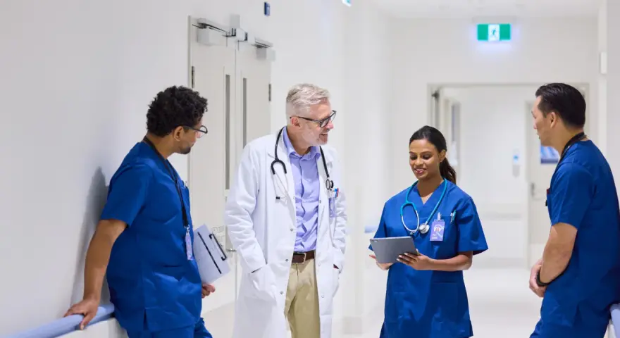 Une équipe médicale discute dans un couloir d'hôpital. Un médecin en blouse blanche échange avec trois infirmiers en uniforme bleu. Ils semblent discuter d'un cas, peut-être en lien avec du infirmier en ligne.